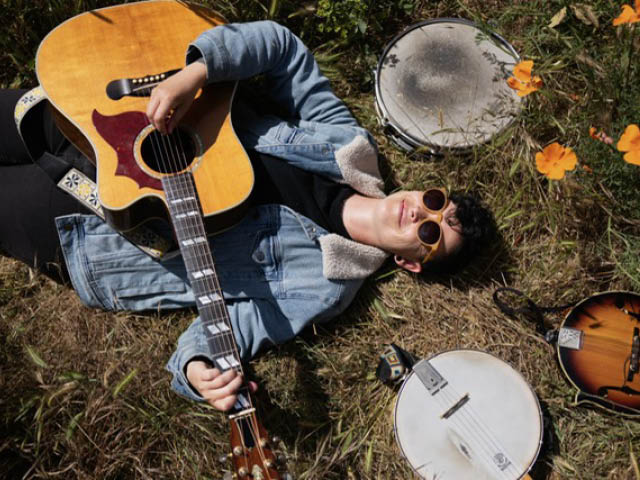Abby Posner, a young white female-presenting musician with a guitar, laying on the grass surrounded by various stringed instruments