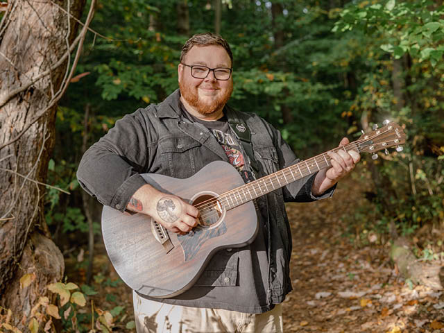 Cam Caruso holding a guitar on a path in the woods