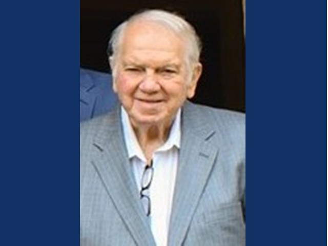 Smiling gentleman in blue-gray suit jacket and light colored button up shirt with glasses tucked from second button.