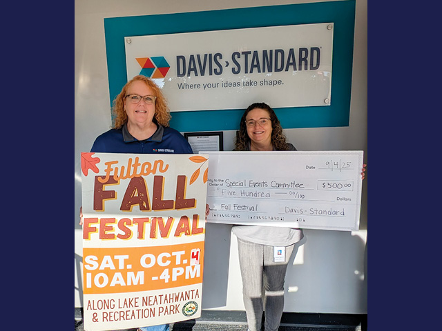 Two women stand in front of a Davis-Standard wall sign, one holding a poster with information about the fall festival and the other holding a presentation check for $500