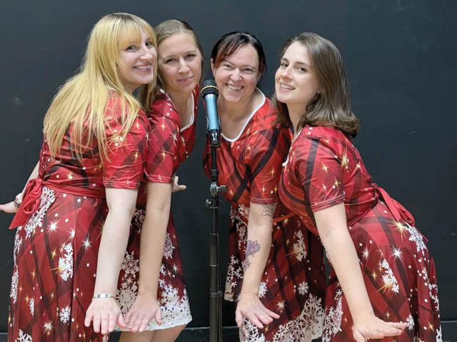 four women in matching holiday dresses standing around a microphone