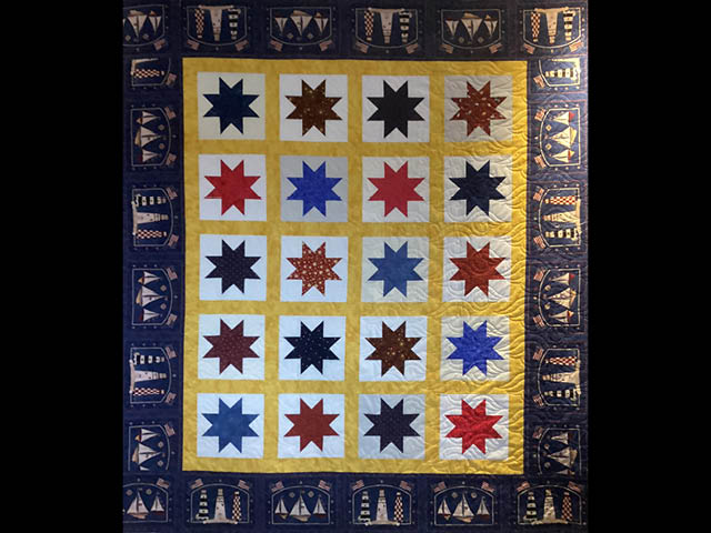 Quilt with starbursts in red, blue, and several dark patterns. A dark border around the edge includes a repeating image of three lighthouses with two American flags waving above them.