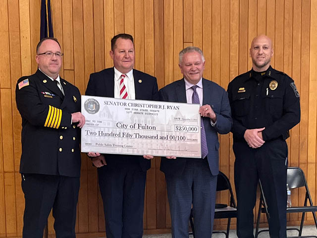 Four white men stand and hold up a ceremonial big check for $250,000