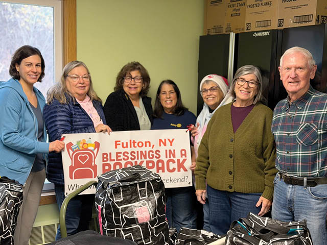 A group of white women and one white man stand in a small room with backpacks and a sign announcing the Fulton Blessings in a Backpack program