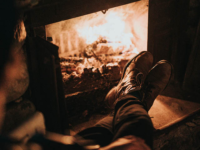 Photo of a dark room with a fireplace in use; resting on the hearth are two feet in a pair of shoes