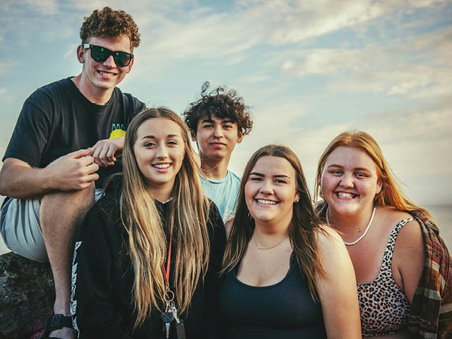 Five teens and young adults smiling and posing outside under a blue sky