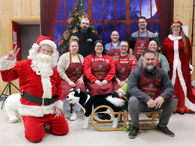 A group of festively dressed volunteers, including Santa kneeling on his knees and giving the camera a peace sign in the front left and Mrs. Claus standing in the back right