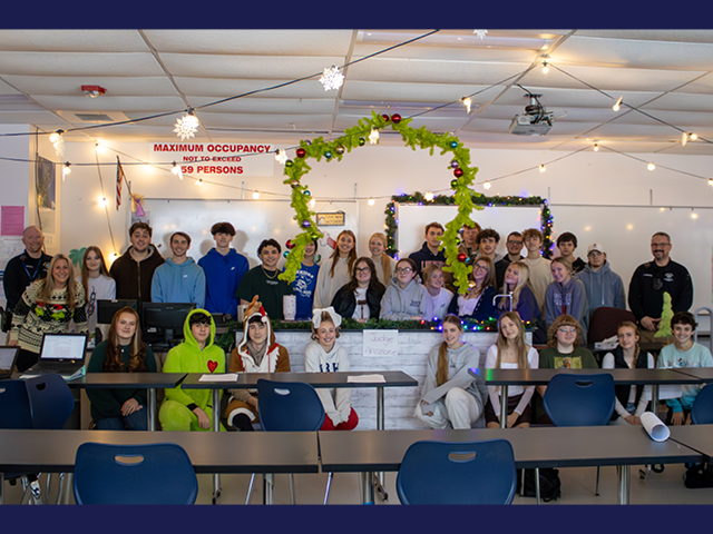 a class posing in the classroom; one of the students is dressed in a green onesy with a heart on it, presumably as the Grinch