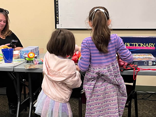 Two young girls from behind as they browse items at a table. A volunteer with Organize Oswego looks on.