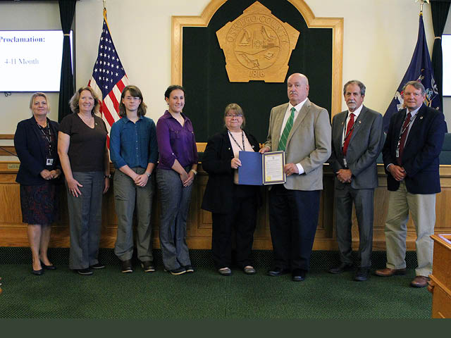 A large group of young and old white people standing in the legislative chambers with a proclamation being presented to the 4H representatives