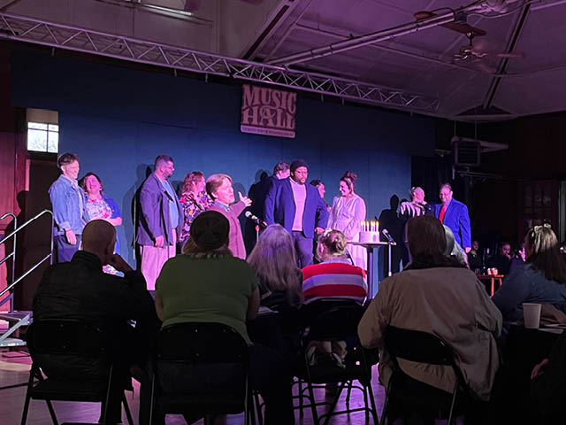 Photo of the full cast of the production of Company on-stage, with audience members visible in the foreground