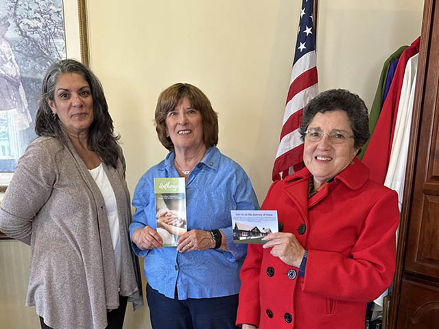 Three women standing in front of an American flag, two of whom are holding up literature about Anthony House