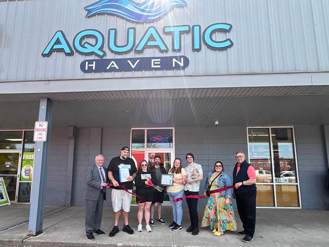 Mayor Jim Rice, Darrick and Amy Venton, Chamber ambassadors and friends and family cutting a ribbon in front of Aquatic Haven's storefront
