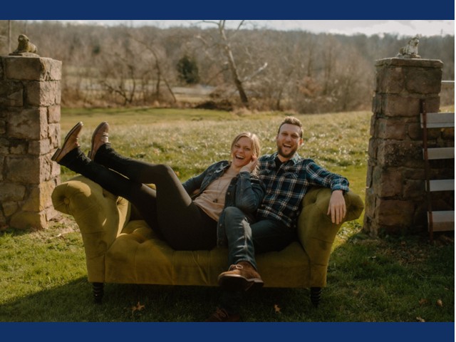 man and woman on green sofa in grass with stone pillars behind them. Man in jeans and blue flannel top sits with legs crossed. Blonde woman with feet on arm of sofa and back against mn wears jeans, pink top and denim jacket.