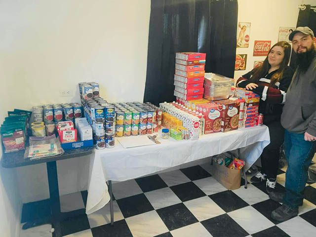 A table filled with meal ingredients; a woman and man stand off to the right side of the table