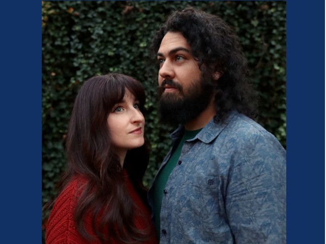 woman with ong dark hair looking towrds man with thick curly shoulder length hair and black beard turned towards woman :greenery in background