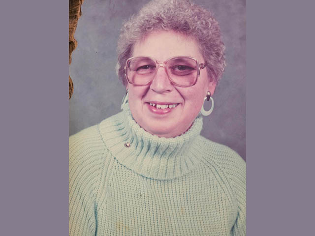 An older woman with tightly curled short gray hair, wearing a green sweater and glasses