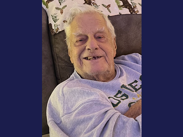 An elderly white man with white hair sitting on a couch arms crossed with a smile on his face