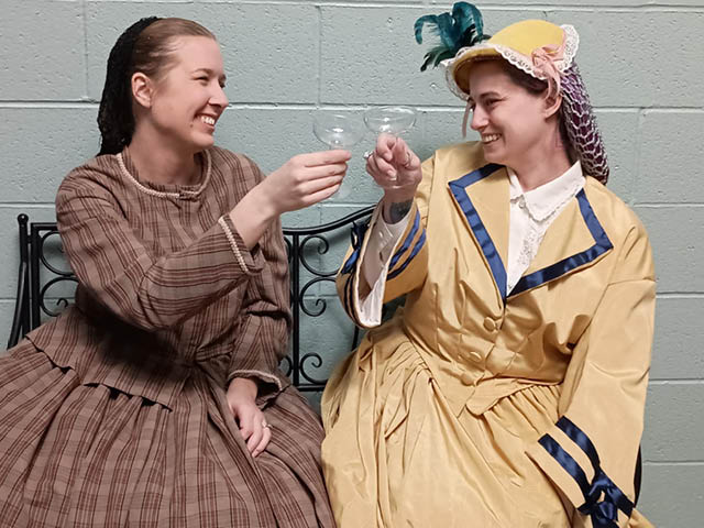 Two women in period costumes toast with empty champagne glasses
