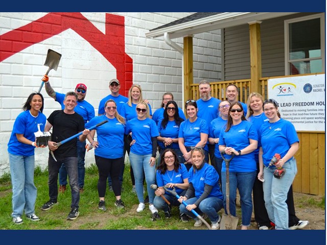 sixteen people standing, two kneeling, some with shovels, wearing blue shirts in front of an image of house on a white wall and a porch with banner on it