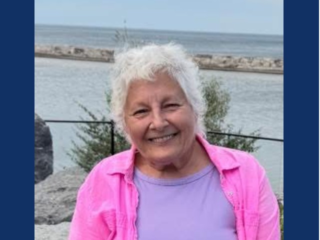 smiling woman with short gray hair in lilac shirt with pink collared shirt over top smiling on beach