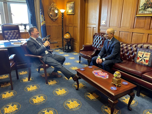 Tow men in suits sitting in an assembly office with leather couch, coffee table and desk  in background. 