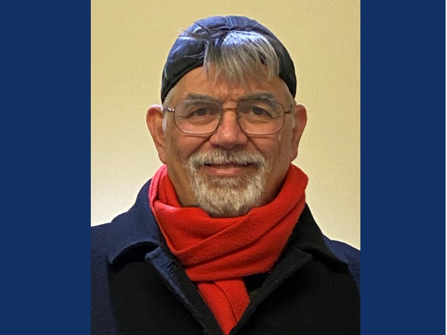 Man with glasses, graying beard, backward baseball cap with bit of hair hanging out tab smiling with blue collared coat and orange scarf wrapped at neck.