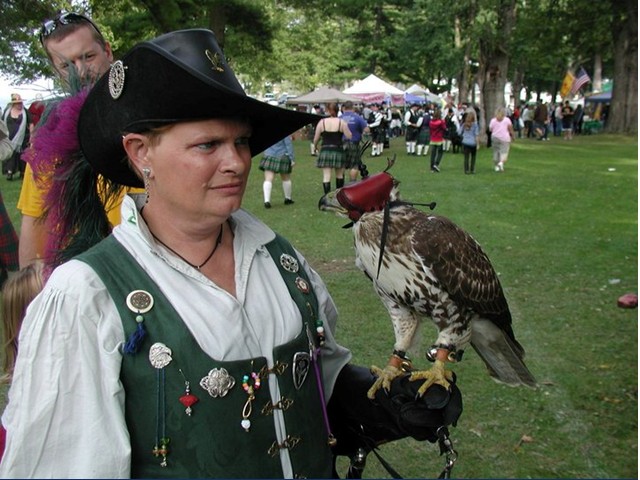 Woman in medieval outfit, hat, eather glove, with capped hawk standing on wrist and hand.