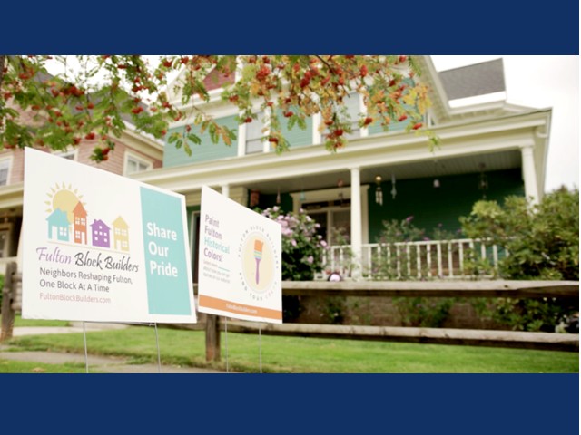 two signs in front of large newly painted home, blue with white trim and white porch, and green grass in front yard