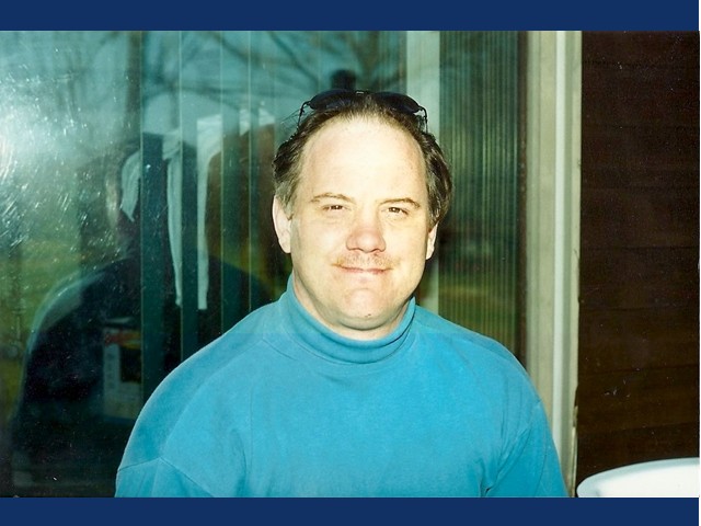 short dark haired grinning man with light blue turtleneck