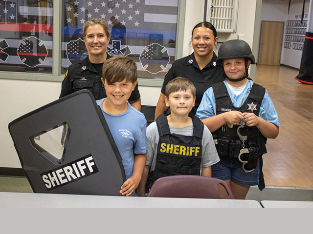 Three students pose with sheriffs; the kids are wearing bullet proof vests and one holds a riot shield