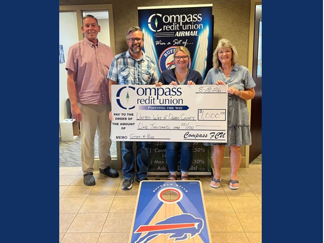 Two men and two women holding a large check in front of compass sign and a buffalo bills corn hole game on floor in front.