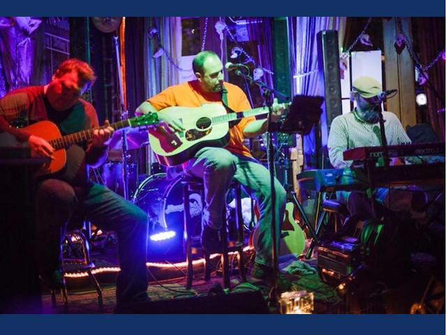 Two men sitting on stage playing gitars and one man sitting behind a keyboard