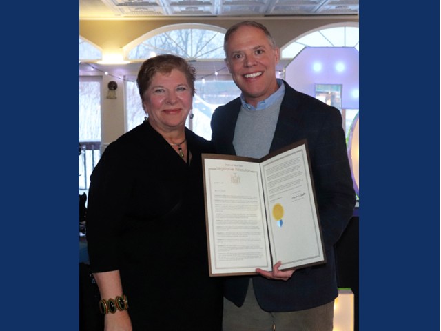 Woman with short dark hair and black top receiving a proclamations from man with blue shirt and dark suit jacket.