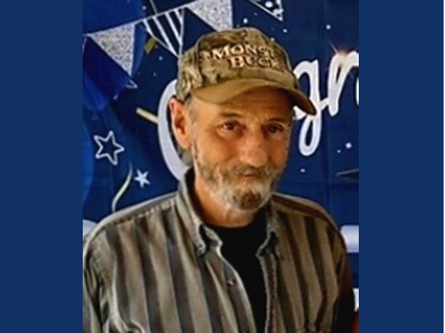 manwith gray/brwon mustache and beard wearing a brown camoflauge baseball cap and brown striped shirt with black t-shirt beneath 