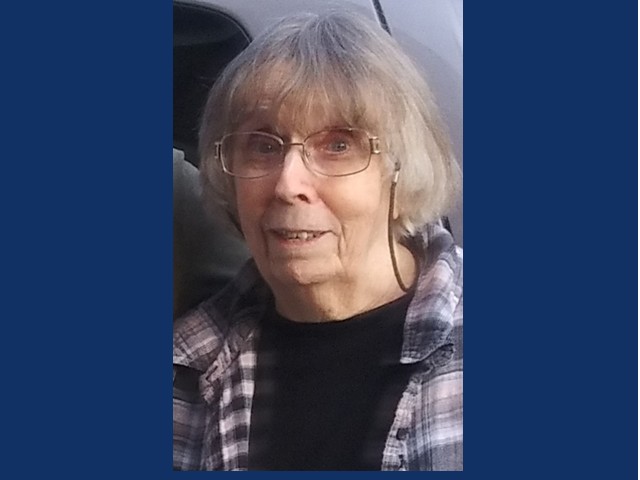 Older woman with gray short hair, glasses wearing a dark shirt with a plaid flannel covering it
