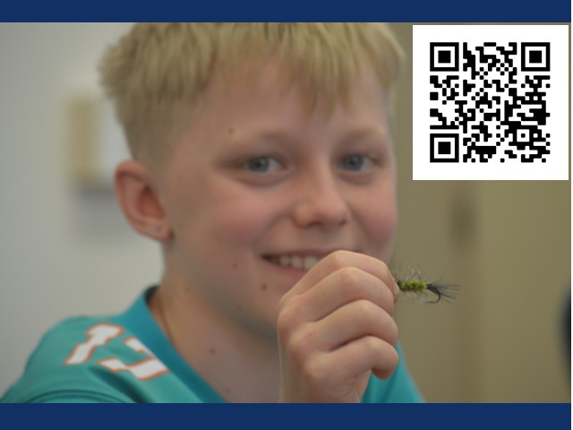 Young blond boy in bluish shirt holding up a fly for fishing.