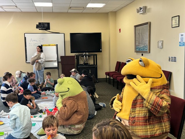 children sitting on floor coloring with big green frog an big yellow frog. Adults scattered within.