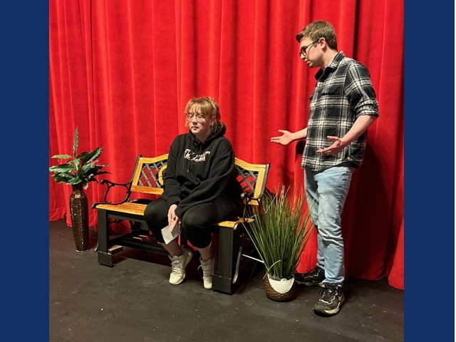 A woman sitting on a park bench with a black hoodie and plants on each side. A young man with his hands pointing up at his hips wearing a flannel shirt and jeans.