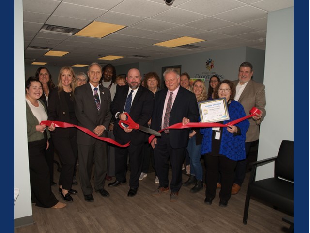 Several men and women holding a long red ribbon across a foyer with a man in middle holding big red scissors after a cut in ribbon.