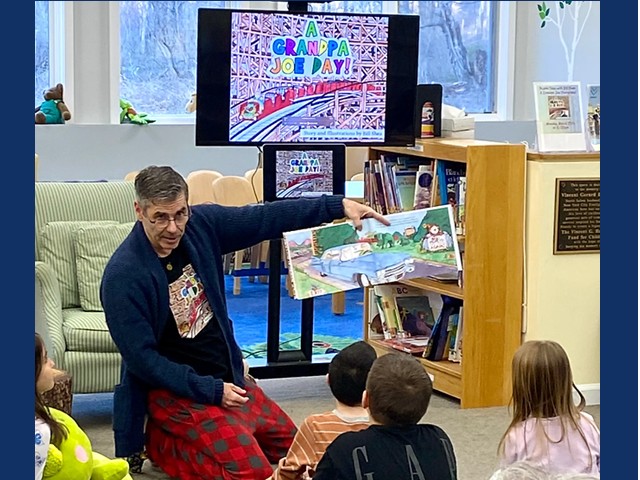 Man with short dark hairsitting on knees on floor holding up book for several kids to see picture, A TV screen in background shows cover of book.