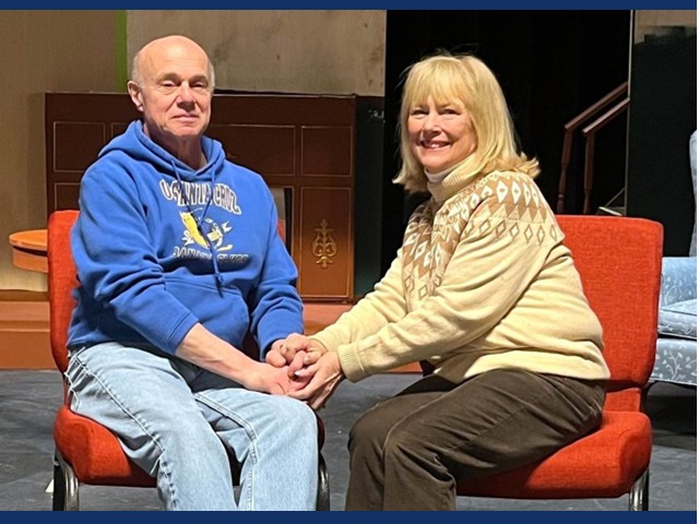 man in blue hoodie and jeans sitting next to a woman with shoulder length hair in sweater and brown pants.