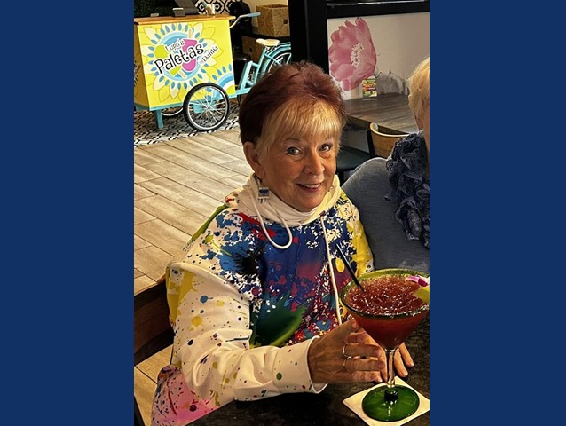Older woman with short dark hair and gray surrounding face smiling with large cocktail in front of her. She wears splattered  blue,and yellow paint on a white top