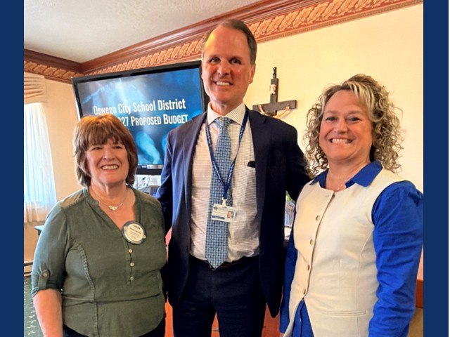tall  man in middle in suit with light blue tie with brown haired woman on left in green shirt and blonde curly haired woman on right with bright blue shirt and white vest over top