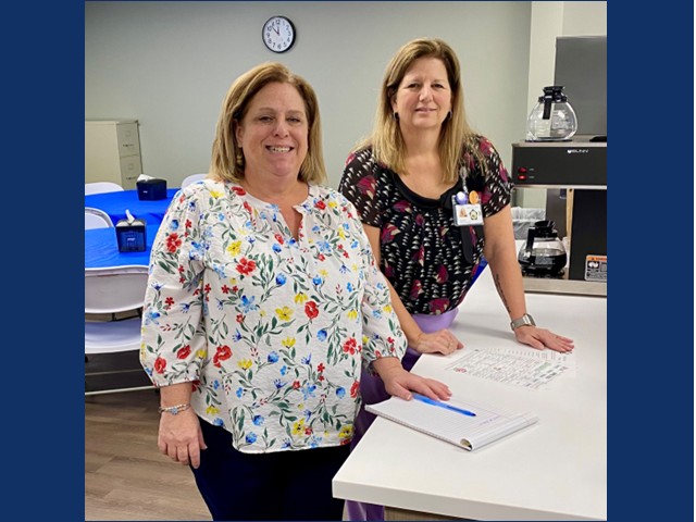 Two women first with shoulder length sandy brown hair in white shirt with red and blue floral pattern, secound with over the shoulder light brown hair in black short sleeved top with floral pattern. Both have hand on table with paper and pen in front of them.