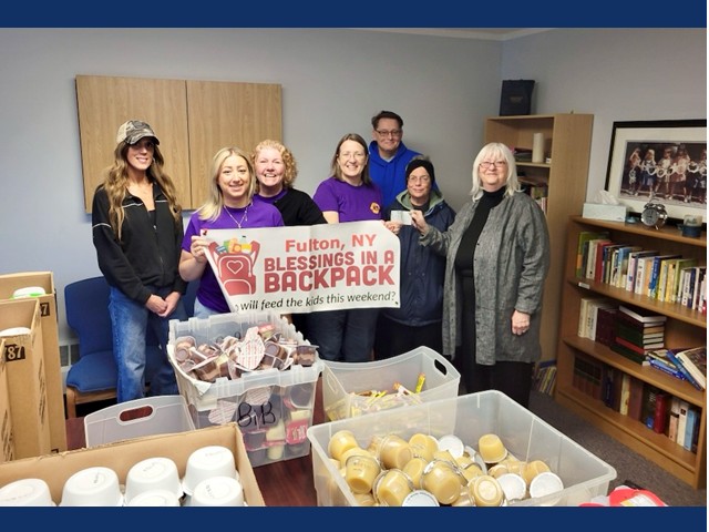 A group of people  holding "blessings in a backpack" banner in fromt of bins and boxes  of nonperishable food