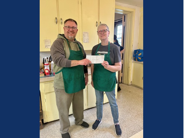 man with green apron, short dark hair, gray short beard, and glasses hands check to short gray haired woman in green apron while standing in front of counter