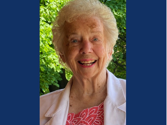 Smiling older woman with full silver hair in white colored blazer over red and white top. Green trees in background.