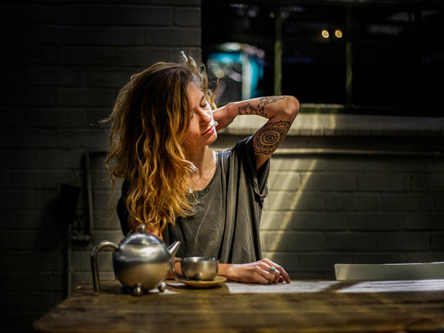 A tattooed white woman sitting at a table and reaching up to rub her neck
