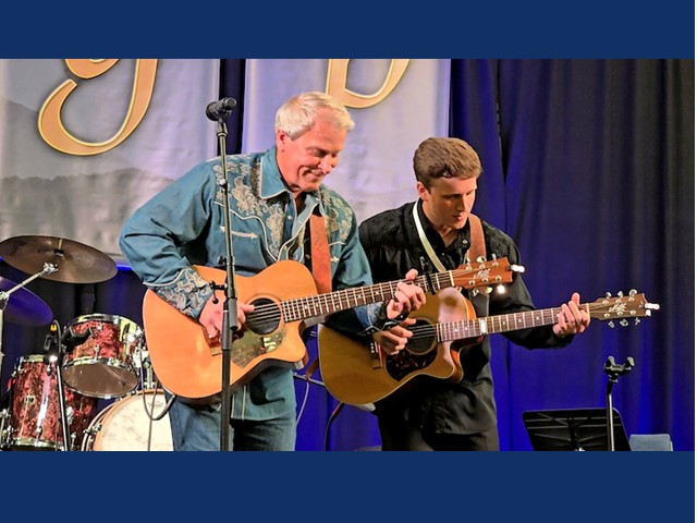 Man with short silver hair, denim shirt, and acoustic guitar playing next to a young dark haired man also playing accoustic guitar